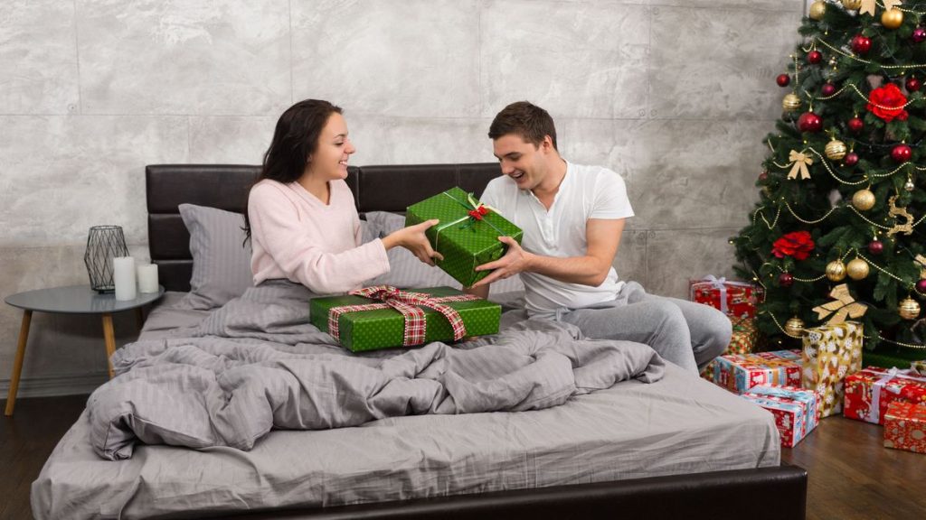 young happy couple in pajamas giving each other presents while sitting on the bed in christmas morning